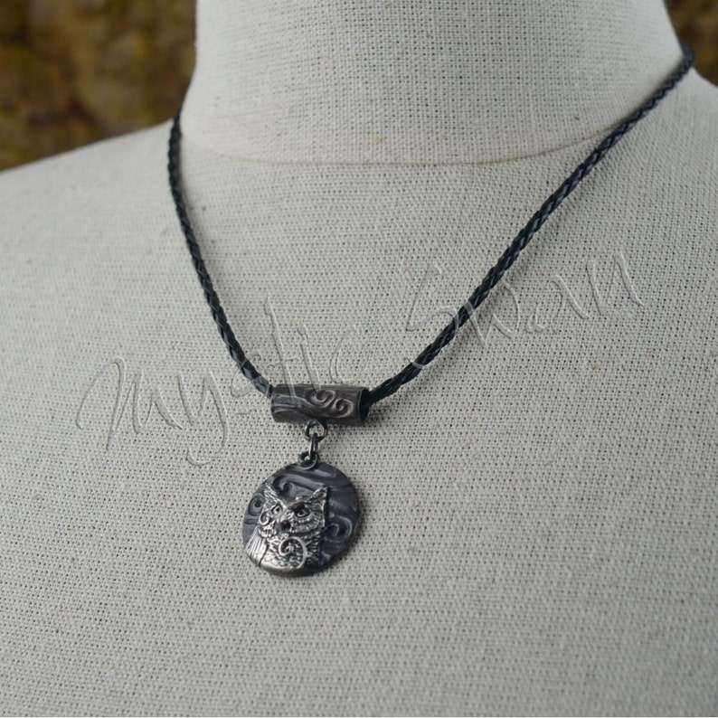 May include: A silver-toned pendant necklace featuring an owl design. The pendant hangs from a black braided cord. The owl is detailed with etched features and is set against a textured background. The necklace has a decorative bar above the pendant.