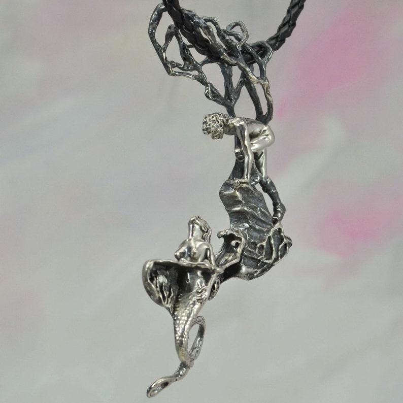 May include: A silver pendant necklace featuring a mermaid and a figure climbing a rocky cliff. The pendant is attached to a black chain.