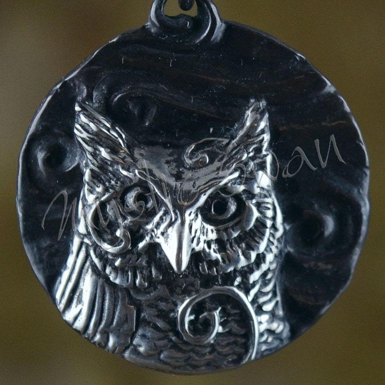 May include: A detailed, silver-toned pendant featuring a sculpted owl design. The owl is centered on a circular backdrop with a textured surface. The pendant has a loop at the top for hanging and is suitable for jewelry.