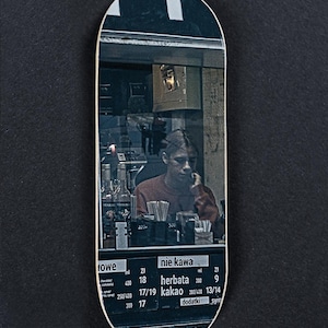 Pode incluir: Um deck de skate com uma fotografia de uma pessoa num café. A pessoa está ao telefone. O deck tem um fundo preto e um menu com preços em polaco.
