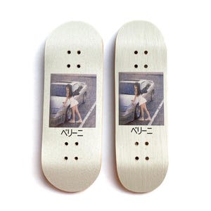 Könnte beinhalten: Zwei Finger-Skateboards aus Holz. Jedes Board hat ein gedrucktes Bild einer Person neben einem Auto. Der Text "ベリーニ" steht unter dem Bild. Jedes Board hat vier Sätze von zwei Löchern.