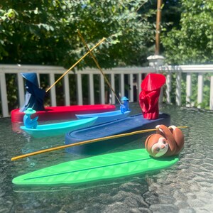 May include: A collection of four incense holders featuring three boat-shaped designs with figures wearing conical hats in red, blue, and light blue, and one green leaf-shaped holder with a brown sloth figure holding a thin incense stick.