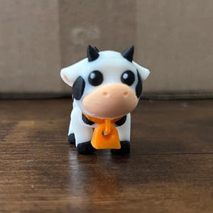 May include: A small white and black cow figurine with orange and black details. The cow is wearing a bell around its neck.