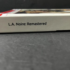 May include: A close-up of a video game box with the title "L.A. Noire: Remastered" printed in black on a white background. The box has a red stripe on the top edge.