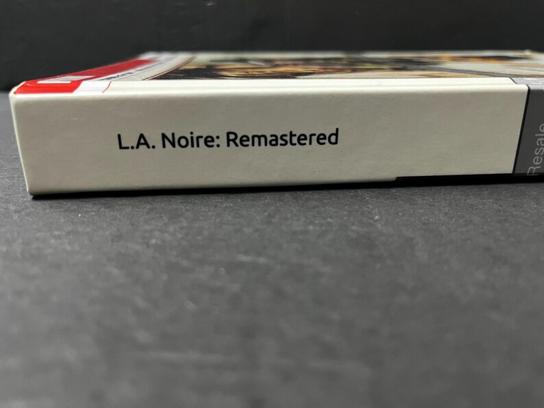 May include: A close-up of a video game box with the title "L.A. Noire: Remastered" printed in black on a white background. The box has a red stripe on the top edge.