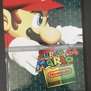 May include: A red, green, and blue Super Mario 64 Nintendo Player's Guide book cover featuring a cartoon image of Mario. The book cover has the text "Super Mario 64" and "Nintendo Player's Guide".