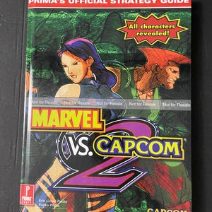 May include: A Prima's Official Strategy Guide book cover for the video game Marvel vs. Capcom 2. The cover features a green, black, and purple background with the game's logo and characters from the game, including a female character with purple hair and a male character with red hair.