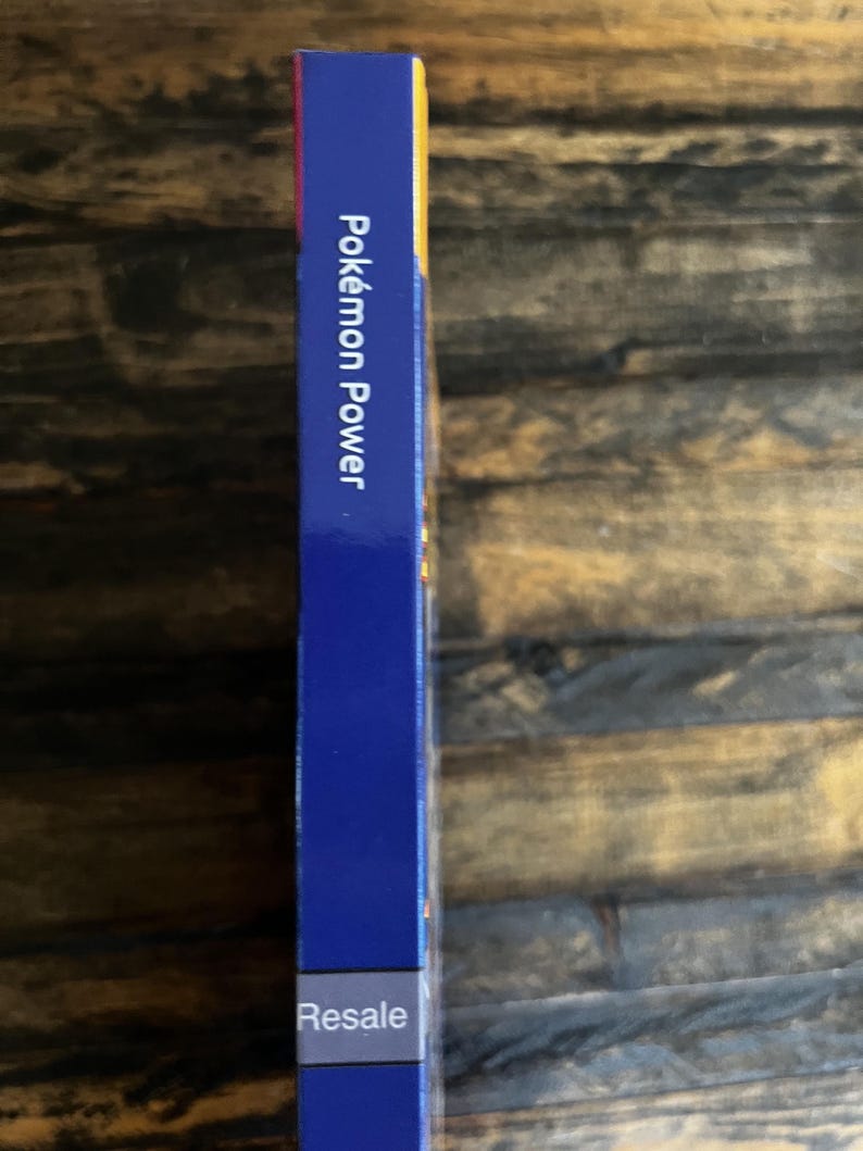May include: A blue rectangular box with the text "Pok&eacute;mon Power" in white. The word "Resale" is printed in white at the bottom of the box. The box is on a wooden surface.