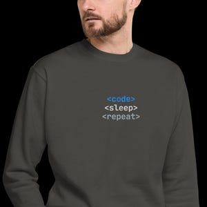 Op de afbeelding: Donkergrijze sweatshirt met de tekst "<code> <sleep> <repeat>" in blauw en grijs. De sweatshirt heeft een ronde hals en lijkt gemaakt van een zacht materiaal. De tekst staat in het midden op de borst.