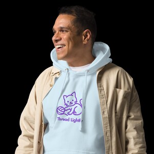 May include: A pale blue hoodie featuring a purple cat design and the words "Thread Light". The hoodie has a drawstring hood and is worn with a tan shirt. The image shows a person smiling and wearing the hoodie.