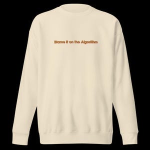 Op de afbeelding: Een crèmekleurig sweatshirt met de tekst "Blame it on the Algorithm" in oranje op de voorkant.