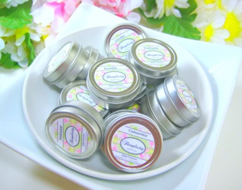 Op de afbeelding: Een verzameling kleine, zilverkleurige blikjes gevuld met een cosmetisch product, gerangschikt in een witte kom. Elk blikje heeft een etiket met een bloemenontwerp en het woord "Simplicity". De etiketten tonen verschillende geurnamen.
