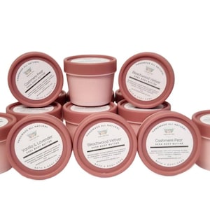 May include: A collection of pink shea body butter containers with brown lids. The labels display the scents: Vanilla & Lavender, Beachwood Vetiver, and Cashmere Pear. The containers are arranged in a stacked display.