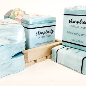 May include: Several bars of artisan soap, labeled "skinplicity," are stacked and arranged on a light background. The soap is a light blue color with a wave-like texture. Each bar is wrapped and labeled with the scent "Whispering Waves" and weighs 4.5 ounces.