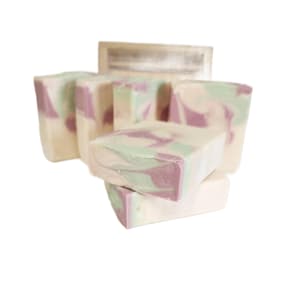 May include: A collection of handmade soap bars with a creamy white base and swirled lavender and mint green accents. The soaps are rectangular and stacked, with a silver-colored label in the background.