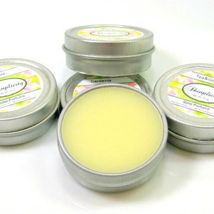 Op de afbeelding: Verschillende kleine, ronde, zilveren blikken met vaste parfum. E&eacute;n blik is open en toont een lichtgele balsem. De etiketten tonen "Gardenia" en "Tea & Ginger" met het merk "Skinplicity".
