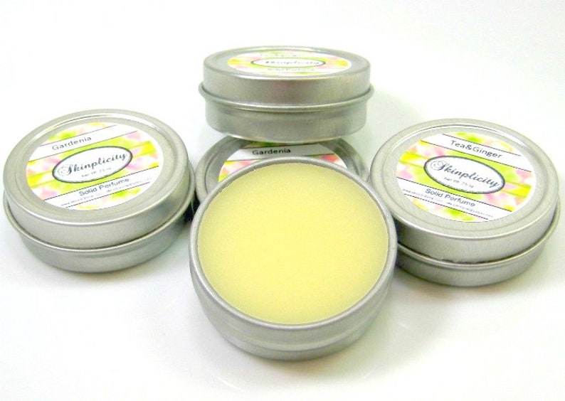 Op de afbeelding: Verschillende kleine, ronde, zilveren blikken met vaste parfum. E&eacute;n blik is open en toont een lichtgele balsem. De etiketten tonen "Gardenia" en "Tea & Ginger" met het merk "Skinplicity".