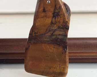 Puede incluir: Un colgante de piedra natural pulida. La piedra muestra una mezcla de tonos marrón, beige y verde oscuro con intrincados patrones de vetas. Un pequeño agujero está perforado en la parte superior para colgar el colgante.