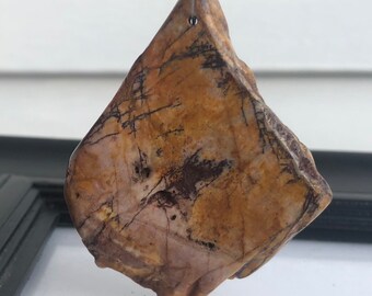 Puede incluir: Colgante de piedra natural de forma irregular, con tonos beige, marrón y negro. La piedra presenta patrones lineales oscuros y está suspendida por un fino alambre plateado, resaltando su textura natural y diseño.