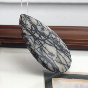 May include: A teardrop-shaped pendant made of stone, featuring a pattern of black veins on a light gray background. The pendant is suspended by a thin, clear string, suggesting it's designed to be worn as jewelry.