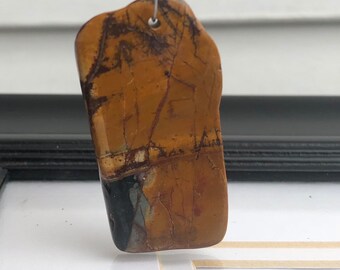Puede incluir: Colgante rectangular pulido, con aspecto de piedra natural. Presenta una mezcla de tonos marrón, beige y negro con vetas intrincadas. Un pequeño agujero perforado en la parte superior para una cadena o cordón.