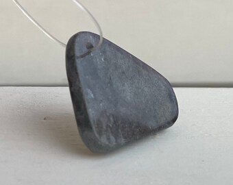 May include: A dark gray, triangular-shaped pendant with a hole at the top for string or cord. The pendant is made of a smooth, polished stone.