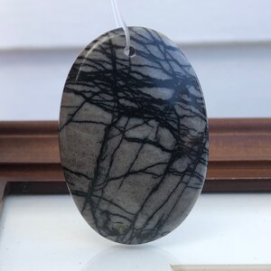 May include: Oval-shaped pendant with a gray base and a striking black vein pattern. The stone is suspended by a thin, clear cord. The pendant's design resembles a map or network, offering a unique and natural aesthetic. The pendant is a jewelry item.