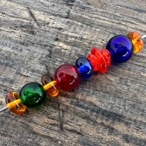 Op de afbeelding: Een streng gekleurde glazen kralen, waaronder groen, oranje, rood, blauw en amber. De kralen zijn geregen aan een draad en zijn gerangschikt in een regenboogpatroon.