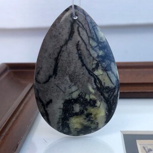 May include: A teardrop-shaped pendant with a gray and black marbled pattern. The stone has a small hole at the top for a string. The pendant is a neutral color and could be worn with many outfits.