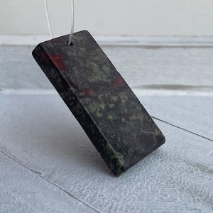 May include: A rectangular pendant made of dark green, black, and red stone with a hole drilled through the top for a cord.