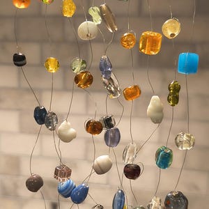 May include: A hanging mobile featuring numerous multicolored glass beads of various shapes and sizes, suspended from a horizontal metal wire by thin, individual silver-toned wires. The beads display a wide spectrum of colors including blue, green, yellow, orange, red, clear, and white, creating a cascading arrangement.