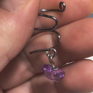 May include: A silver-colored, wire-wrapped ear cuff with a dangling, faceted, purple crystal. The ear cuff has a unique, curved design with a small loop at the top and a hook at the bottom for securing to the ear.