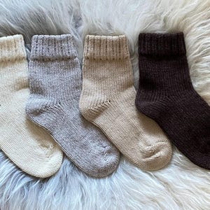May include: Four pairs of knitted socks in cream, grey, beige, and dark brown. The socks are folded over at the cuff and are displayed on a fluffy, white and grey textured surface.