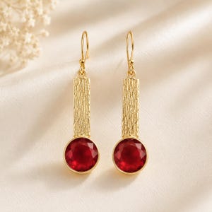 May include: A pair of gold drop earrings featuring a rectangular bar with a textured, hammered surface finish. Each earring is anchored by a round, faceted deep red gemstone set in a smooth gold bezel, suspended from a simple gold hook wire.
