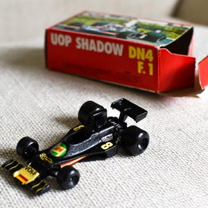 Puede incluir: Un coche de carreras de juguete negro y amarillo con el número 8 en el lateral y la palabra "WOLF" en el alerón delantero. El coche está junto a una caja roja con las palabras "UOP SHADOW DN4 F.1" impresas.