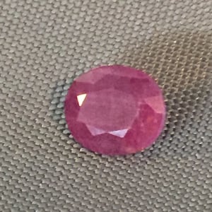 May include: A deep pink gemstone with a faceted cut. The gemstone is round and has a shiny, polished finish.