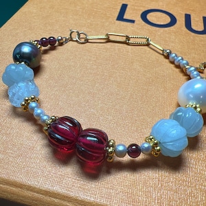 May include: A gold chain bracelet with a variety of beads, including white, light blue, dark blue, red, and purple beads. The bracelet also features a large, white pearl.