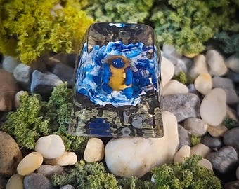 May include: A blue and yellow resin keycap with a small, blue and yellow creature inside. The keycap is surrounded by small rocks and moss.