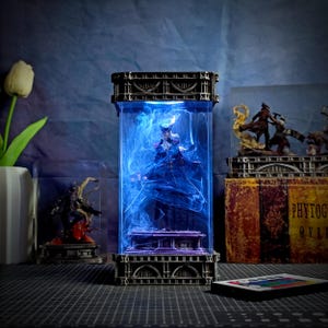May include: A detailed figurine encased in a clear, rectangular display case with a dark, ornate frame. The interior is illuminated with blue light, creating a swirling effect around the figure. Other figurines and a remote control are also visible.