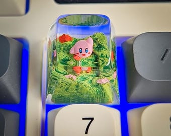 Puede incluir: Un personaje rosa y blanco con ojos grandes está encerrado en un keycap de resina transparente. El keycap está rodeado de resina verde y blanca, que se asemeja a un paisaje herboso.