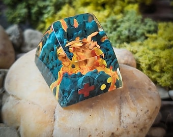 May include: A resin keycap with a blue, orange, and yellow design featuring a dragon. The keycap has a red and yellow cross and two yellow dots.