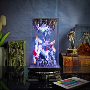 May include: A detailed model of a robot warrior, encased in a clear display box with LED lighting. The robot features a colorful design with red, blue, white, and orange accents. The display includes a rocky terrain base and a remote control for the lights.