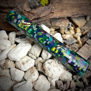 Puede incluir: Un bloque de resina para bolígrafo colorido y multicolor con motas verdes, azules, amarillas, rosas y blancas. El bloque es cilíndrico y tiene un acabado liso y brillante.