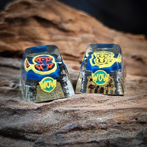 Puede incluir: Dos teclas de teclado personalizadas con un diseño azul, amarillo y rojo. Una tecla tiene la palabra "WOW" y un diseño estilizado. La otra tecla tiene la palabra "WOW" y un diseño de tigre. Las teclas están sobre una superficie de madera.