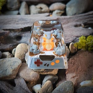 May include: A clear resin keycap featuring a small, light grey and orange figurine. The keycap has a grey and orange design on the bottom, resembling a video game controller.