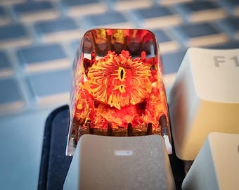 May include: A close-up of a custom keyboard keycap with a red and yellow resin design. The keycap is shaped like a square and has a clear resin top. The design inside the resin is abstract and resembles a flower or eye.