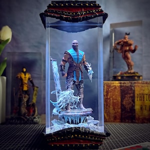 May include: A detailed action figure of a character from Mortal Kombat, encased in a clear display box. The figure is in a fighting pose, with a blue and brown outfit, and is set on a snowy base. The display box has a decorative top.