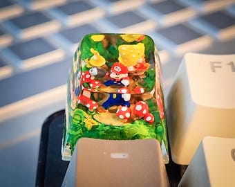 Puede incluir: Un tapón de tecla de resina transparente con una escena en miniatura de Mario y Luigi en un bosque verde con setas rojas y blancas.