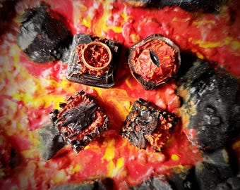 May include: A close-up of four fantasy-themed resin pieces, including a ring, an eye, and two other textured shapes, set against a vibrant red, orange, and yellow background. The pieces have a fiery, volcanic aesthetic with intricate details.