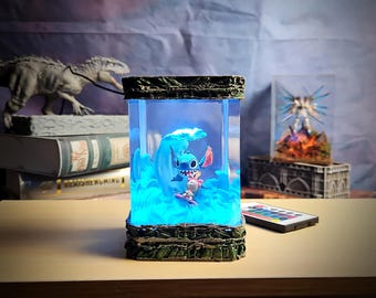 Puede incluir: Lámpara rectangular iluminada con una escena en tonos azules de Stitch surfeando una ola. La lámpara tiene una base y parte superior verde oscuro texturizada, con un mando a distancia cerca. Una figura de dinosaurio y libros están en el fondo.